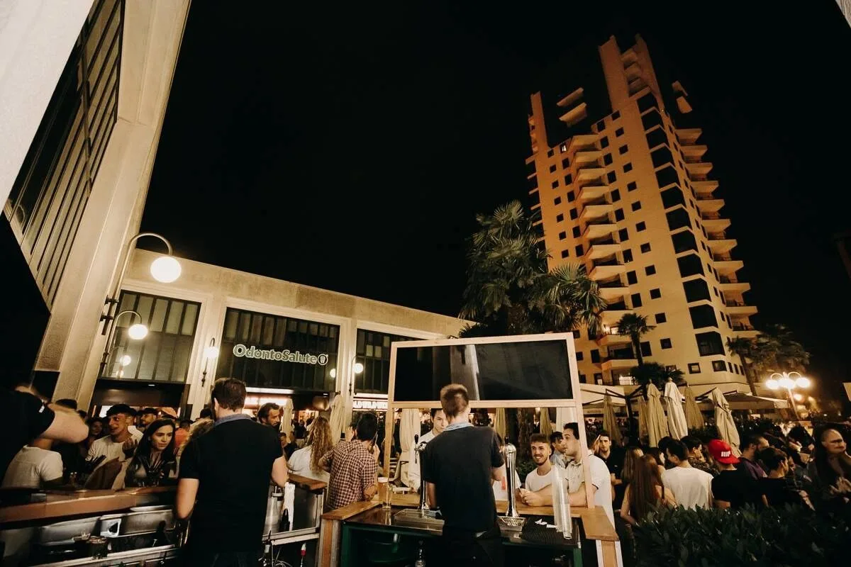 Evento serale all’aperto con folla davanti al Totem Plaza, palme e grattacielo illuminato nella piazza Torri Bianche a Vimercate