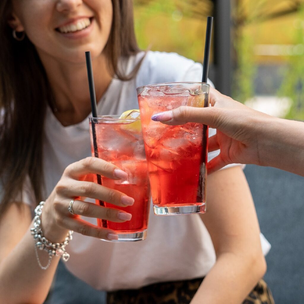 Due bicchieri di cocktail rosso con ghiaccio e fetta di limone, brindisi tra da due ragazze sorridenti sedute all’aperto durante un aperitivo.