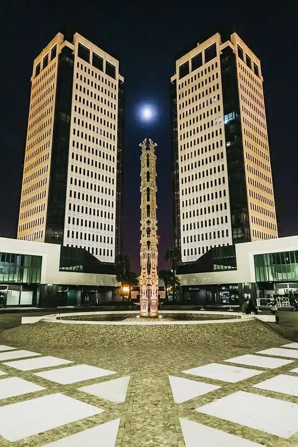 Due grattacieli gemelli illuminati di notte con totem scultoreo centrale e pavimentazione geometrica, nella moderna piazza Torri Bianche di Vimercate