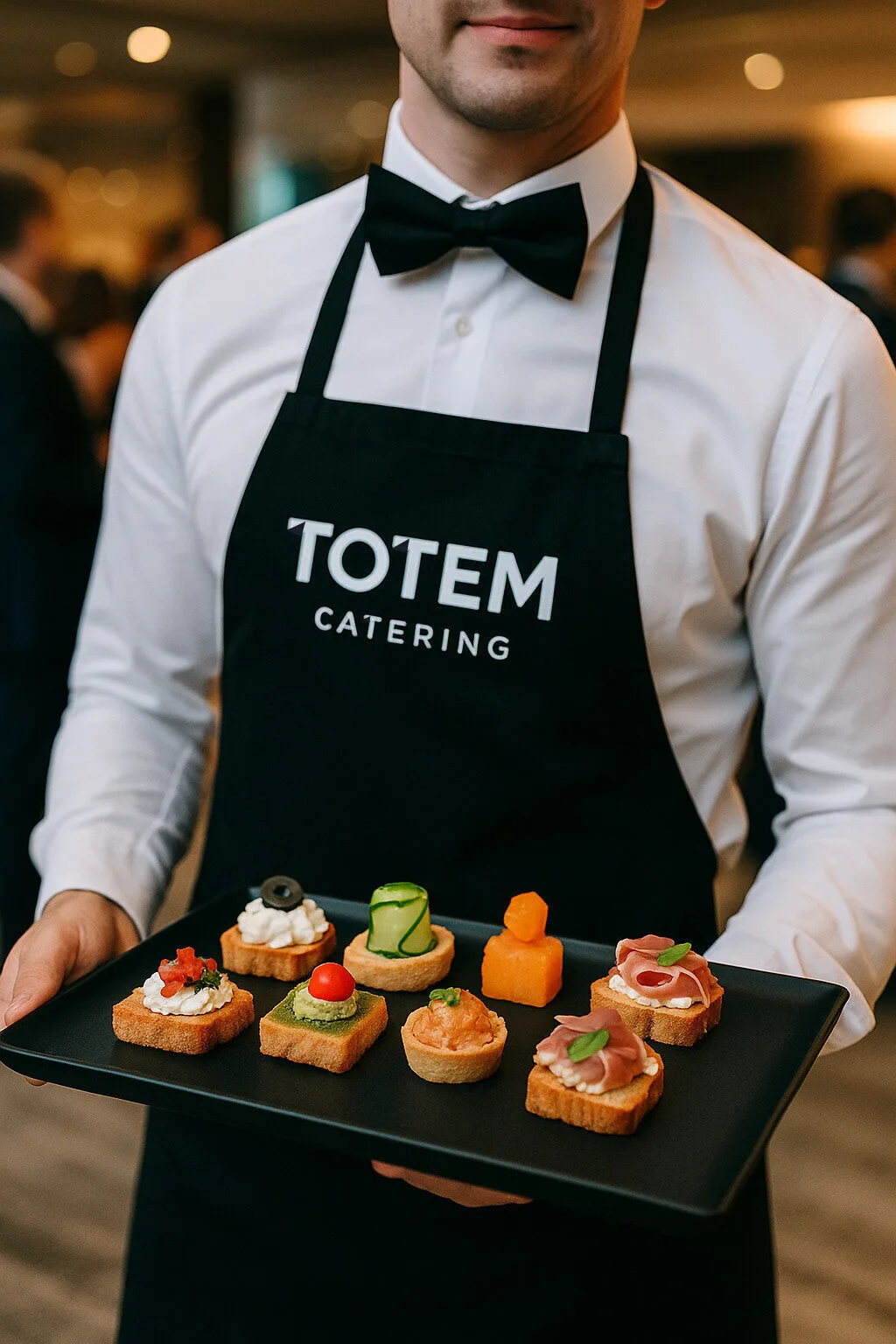 Cameriere elegante con grembiule nero Totem Catering, che porta un vassoio di finger food assortiti tra cui tartine con prosciutto, mousse e verdure, durante un evento raffinato